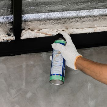 a person wears a glove and sprays foam into a crack along a wall in their house