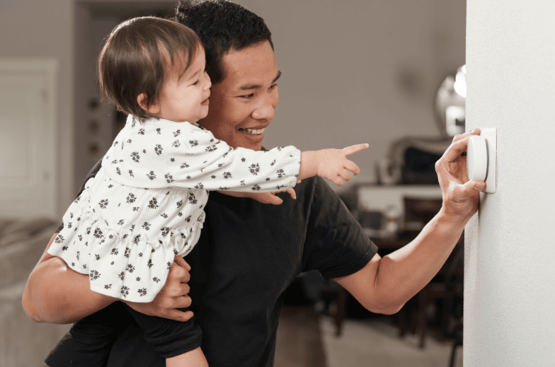 A man holds his baby daughter while adjusting the smart thermostat in his home